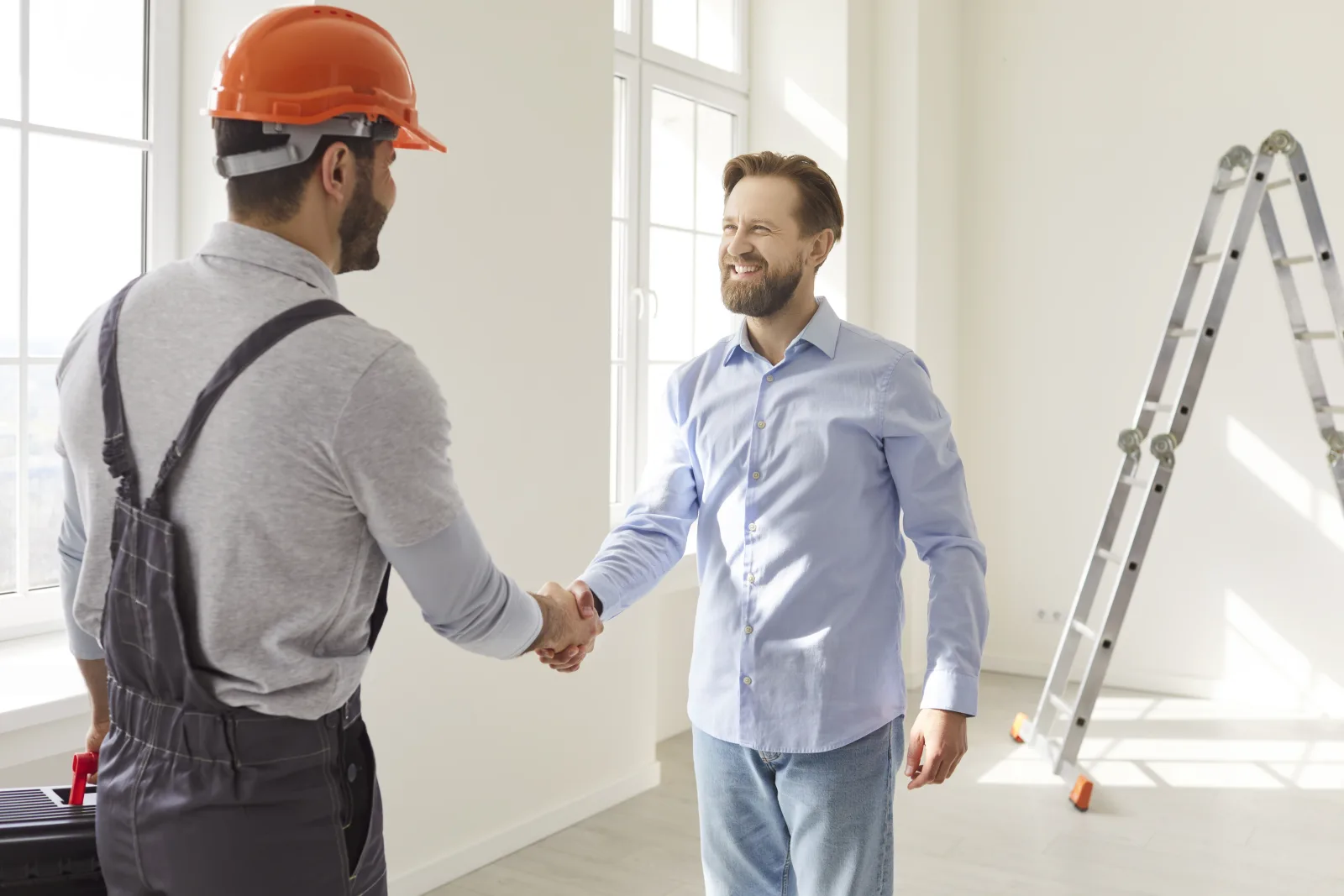 A contractor in a hard hat and a man in a work shirt shaking hands in an empty, freshly finished room, a toolbox at their feet and a folding ladder against the wall — fix and flip insurance in Virginia