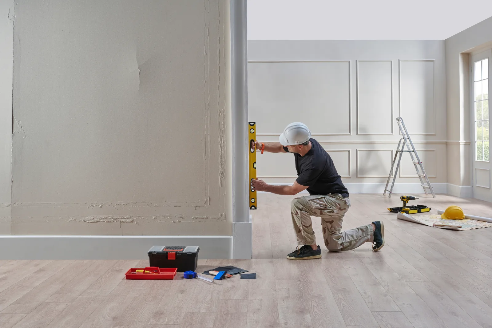 A worker in a hard hat kneeling to hold a spirit level against a freshly skim-coated wall, a stepladder, an open toolbox and rolled plans on the floor behind him — fix and flip insurance in Florida