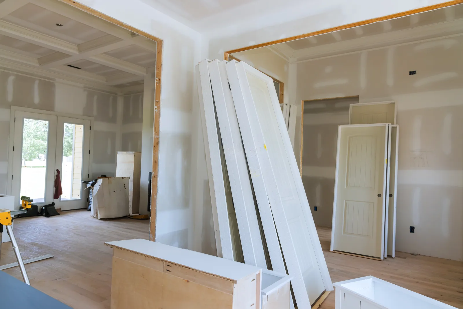 Interior doors and trim stacked against taped drywall in a house at trim-out stage, cabinet carcasses on the bare subfloor and daylight through the rear doors — fix and flip insurance in Vermont