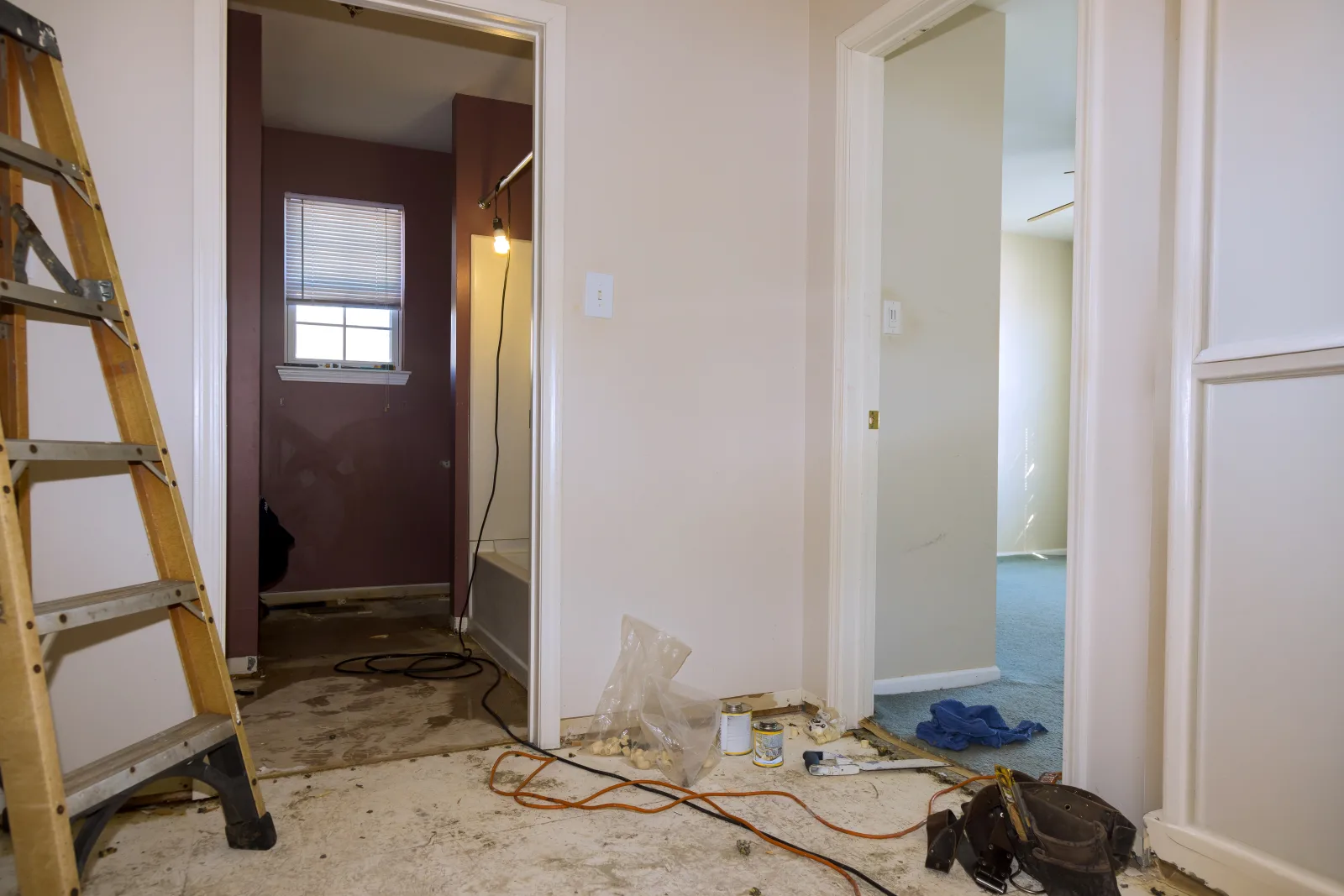 A hallway mid-renovation with the floor stripped back to adhesive residue, a work light strung into the bathroom beyond and a tool belt left on the boards — fix and flip insurance in Georgia