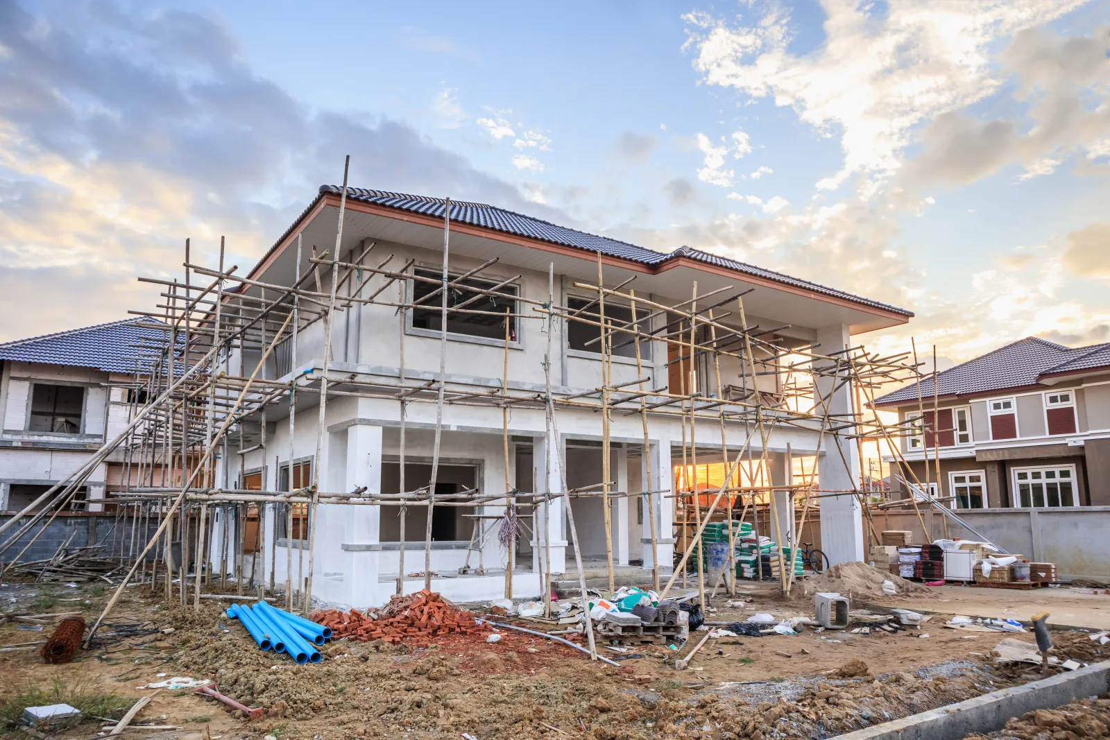 A house under construction behind bamboo scaffolding, its tile roof finished and its window openings still unglazed, with stacked brick and loose pipe on the bare ground — fix and flip insurance in Illinois