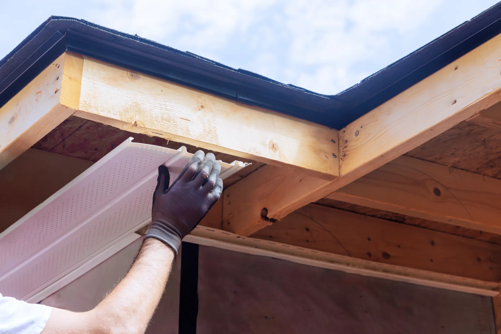 A gloved hand fitting a perforated vinyl soffit panel beneath new fascia and rafter tails, seen from below against the sky — fix and flip insurance in Washington