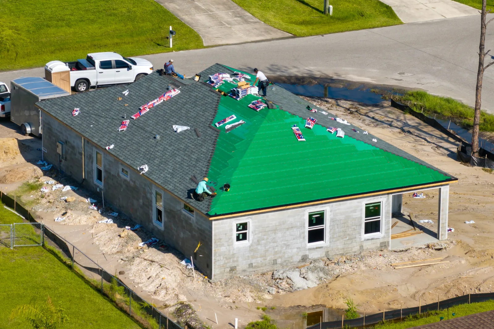 An elevated view of a low block house being roofed, half the slope still bright underlayment and half laid to shingle, with bundles staged across the deck — fix and flip insurance in Arkansas