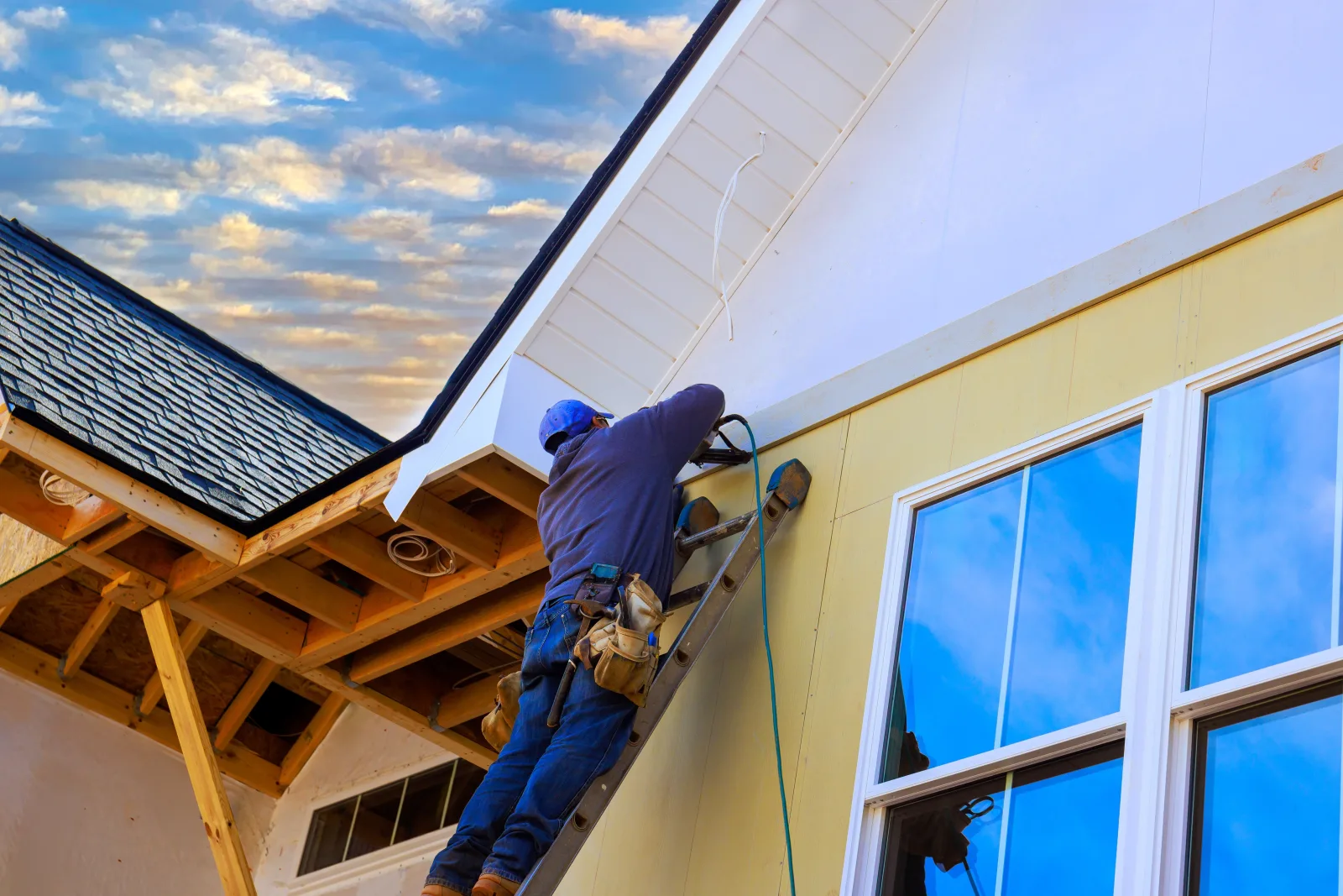 A worker on a ladder fitting trim beneath a gable, exposed rafter tails and sheathing to one side and new windows set in the wall to the other — fix and flip insurance in Tennessee