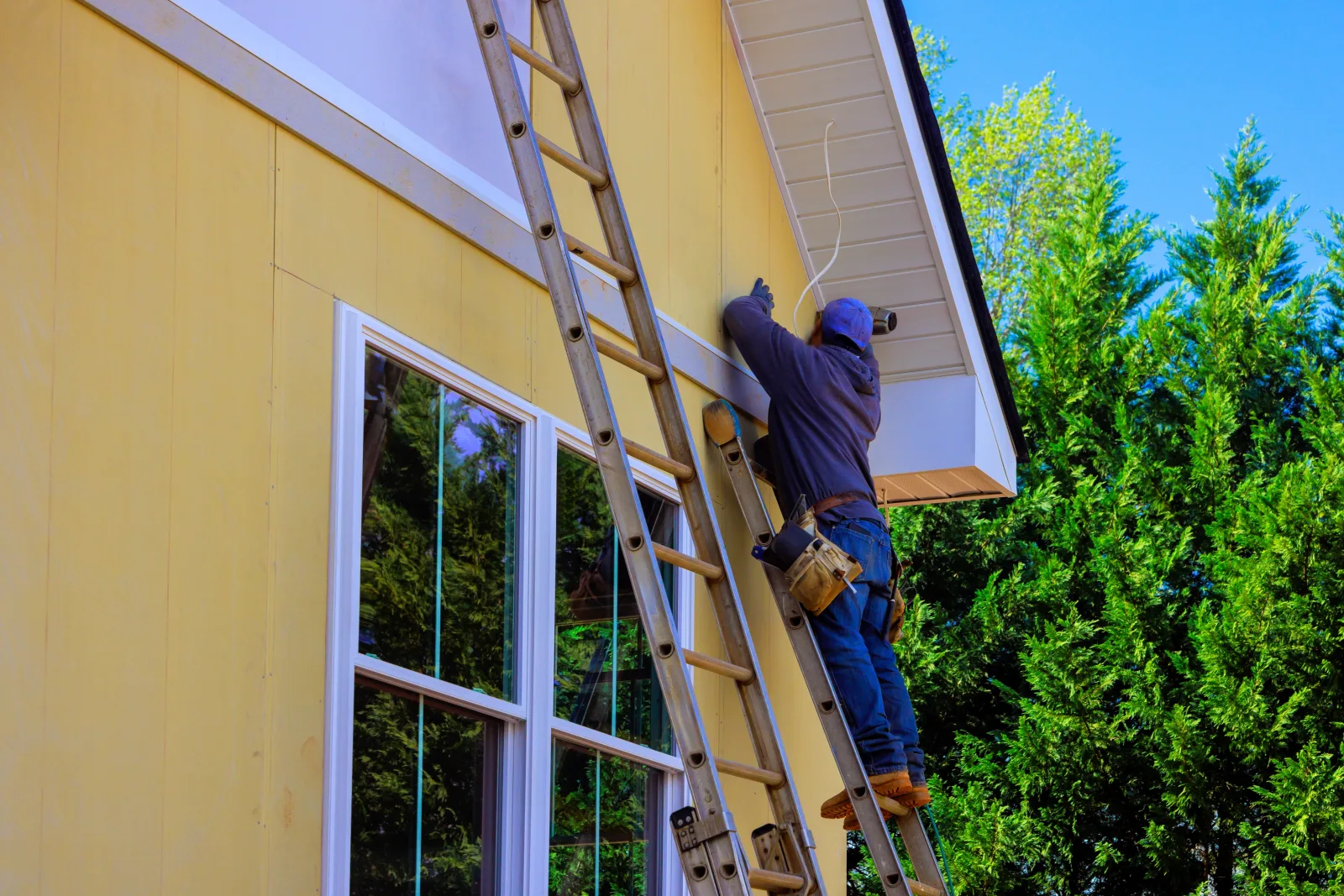 A worker on an extension ladder reaching into the eave of a gable end, the wall below sheathed and the window trimmed out — fix and flip insurance in Oklahoma