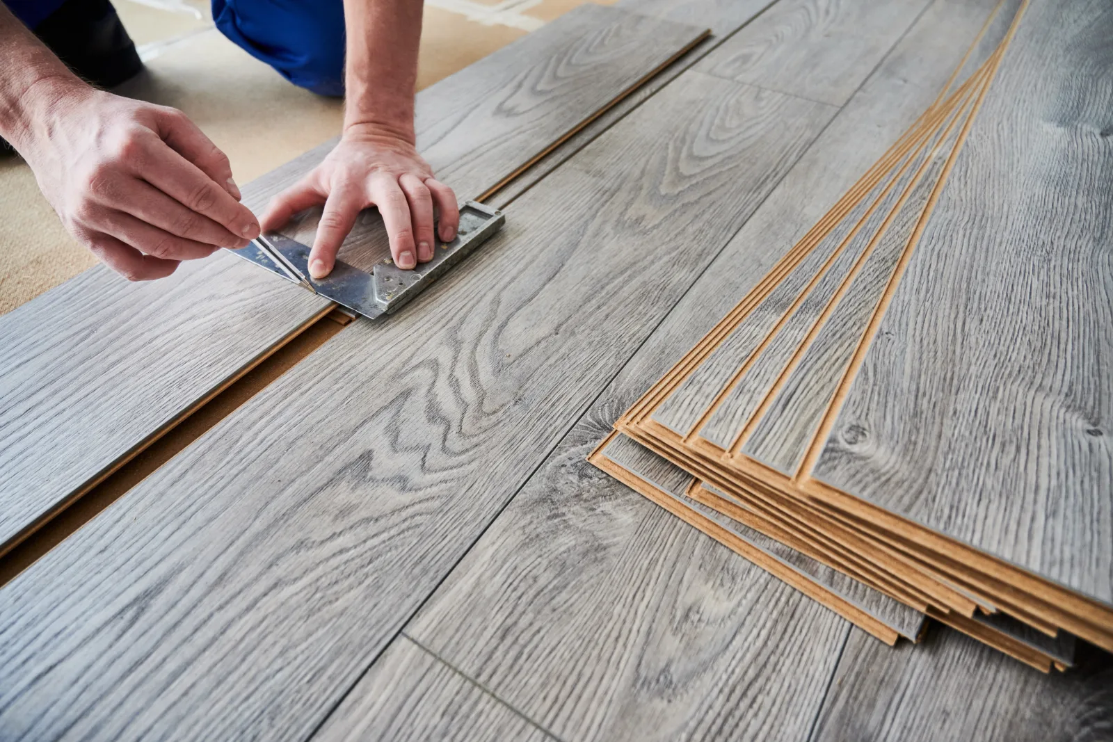 Hands marking a gray wood-look laminate plank with a metal square, a fanned stack of tongue-and-groove boards waiting beside the run already laid — fix and flip insurance in Ohio