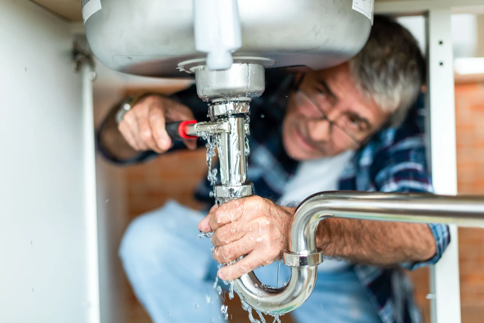 A man working on the chrome trap beneath a stainless sink while water runs from the open joint over his hand — fix and flip insurance in Nebraska